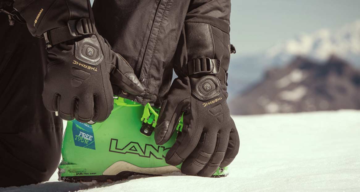 Auf Schnee kniend zu sehende Person, die grüne Skischuhe mit schwarzen Thermic-Handschuhen anfasst.