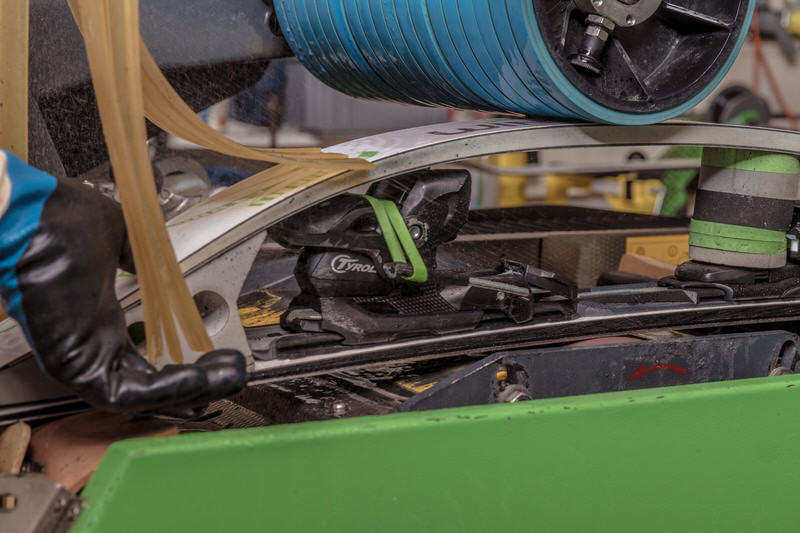 Welche Struktur eignet sich wofür? Und welche ist für einen sportlichen Allround-Pisten- Fahrer? Eine Skibindung wird unter einer blauen Walze auf einer grünen Schleifmaschine bearbeitet. Eine behandschuhte Hand führt den Vorgang.