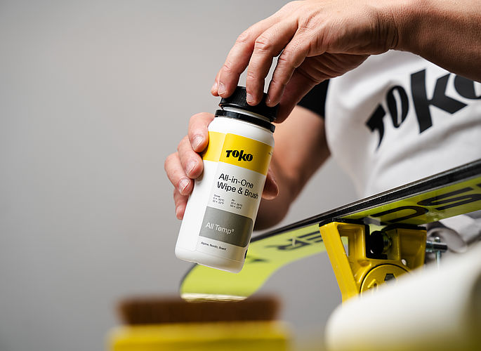 Perfekte Ski- und Snowboardpflege mit dem neuen Toko All-in-One Wipe & Brush Kit Eine Hand öffnet eine Flasche "TOKO All-in-One Wipe & Brush" über einem Ski. Der Ski liegt auf einer gelben Vorrichtung.