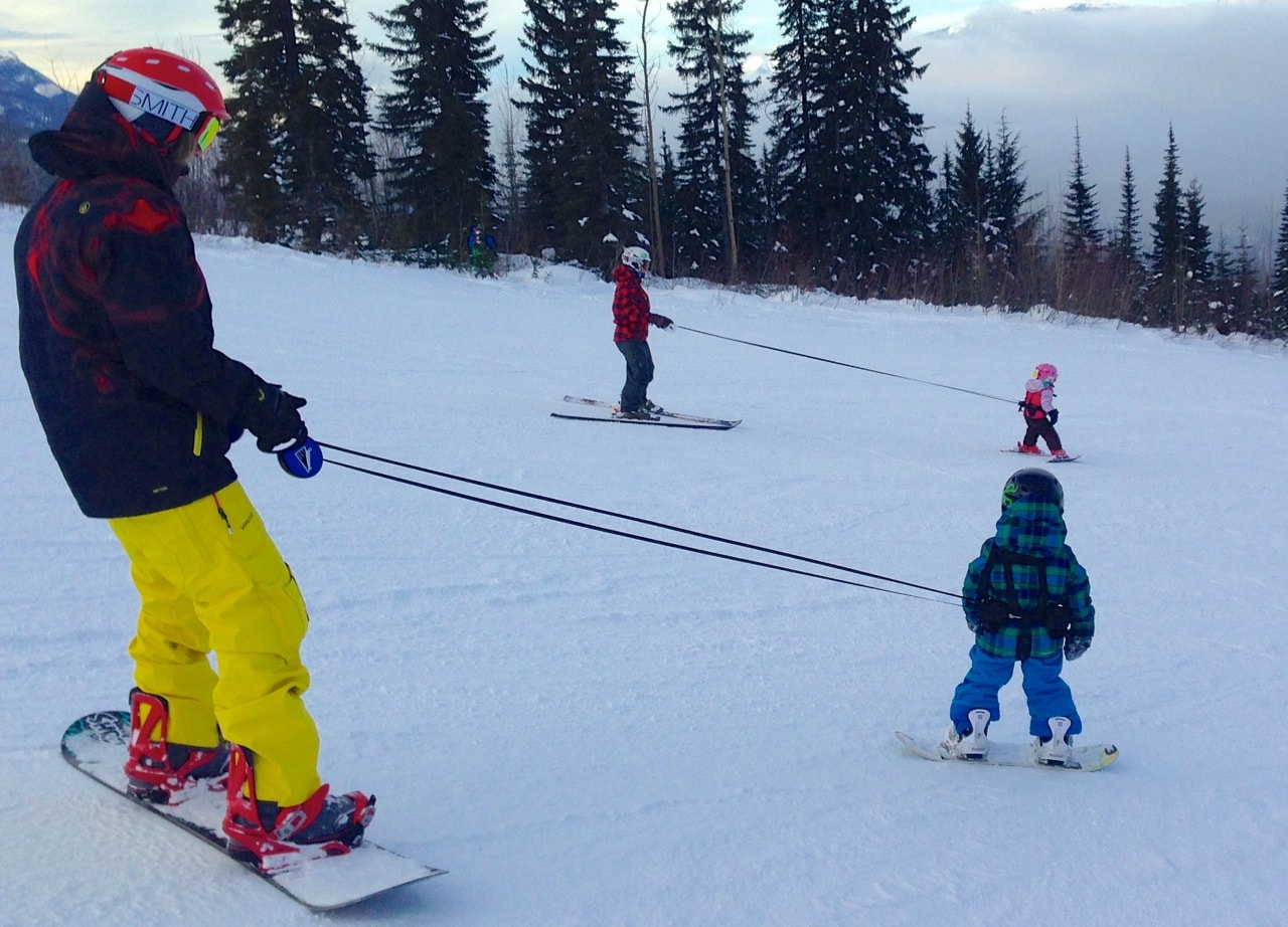Snowboarder Hilfsaktion Auf Schnee: Erwachsener Snowboarder mit Lil' Ripper Gripper Leinen, die Kinder beim Skifahren lernen unterstützen.