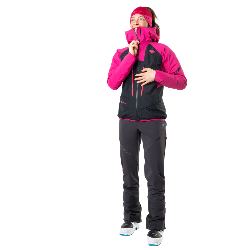 Frau in Dynafit TLT Touring DST Hose, schwarz, pinke Jacke, Skischuhe, Blick nach oben.