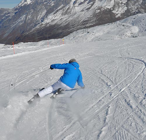 Skifahrer in blauer Jacke und weißer Hose auf verschneiter Piste, im Hintergrund schneebedeckte Berge.