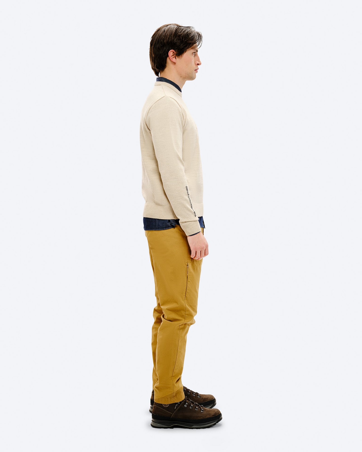 Mann im K-3 ST Ski Sweater Ein Mann, von der Seite, trägt den K-3 ST Ski Sweater in Beige, kombiniert mit braunen Wanderschuhen und gelben Hosen.