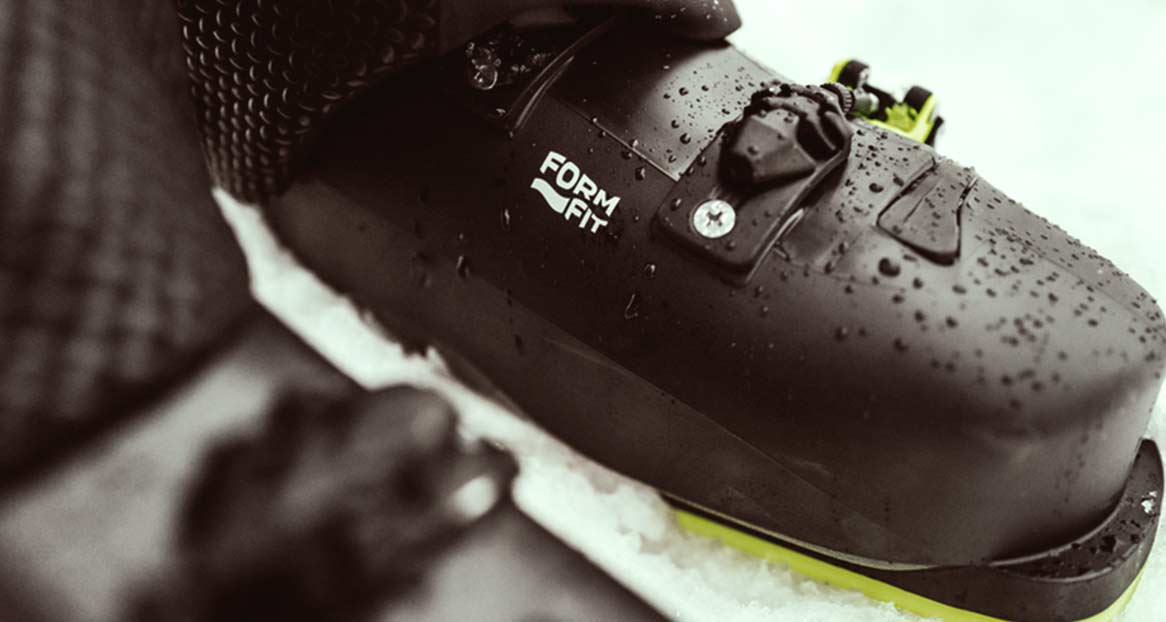 Nahaufnahme eines dunklen, tropfenden Skischuhs mit gelbem Rand auf Schnee. Aufschrift "FORM FIT" ist zu sehen.
