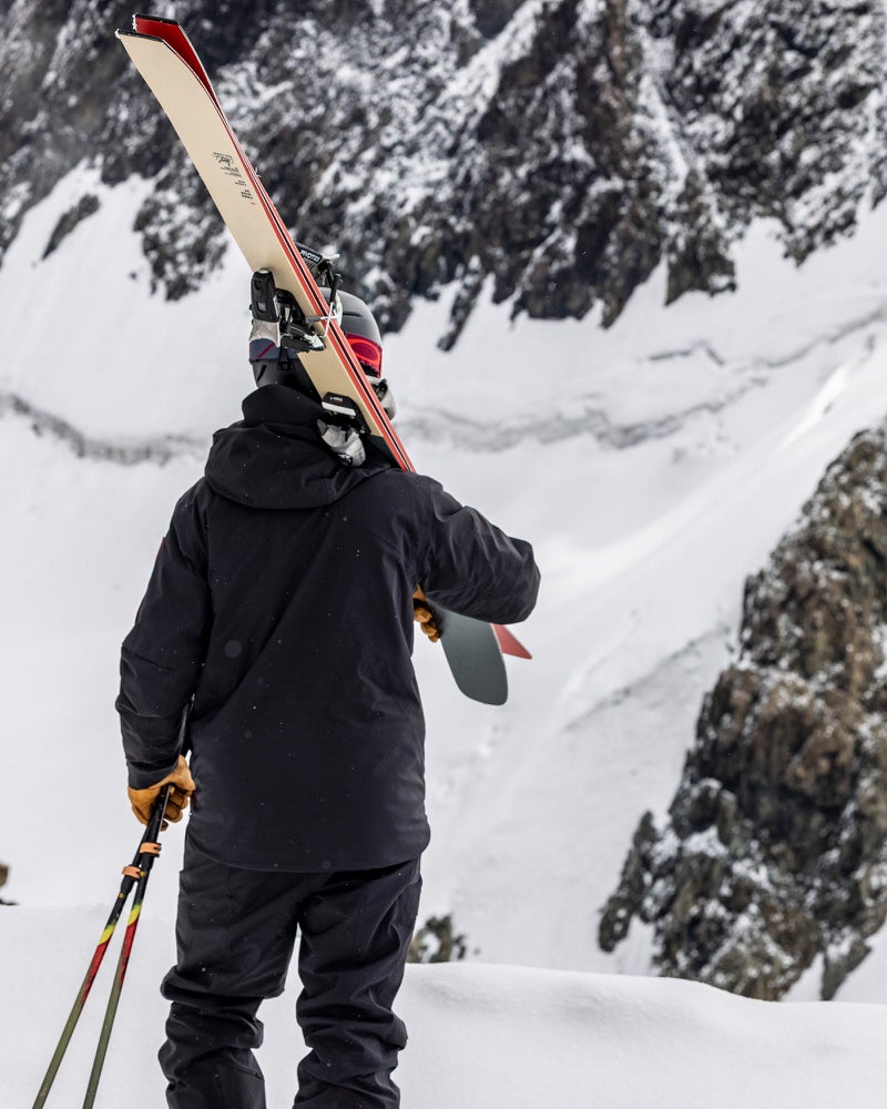 Skifahrer vor Berg Person mit Skiern und Stöcken vor verschneitem Berg, trägt schwarze S-1 Gore Tex Jacke.