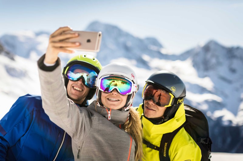 Sicherheit – gut geschützt auf die Piste Drei Skifahrer mit Helmen und Brillen machen ein Selfie vor schneebedeckten Bergen.