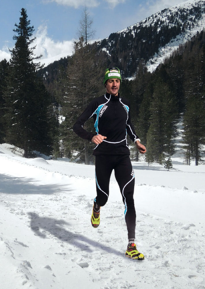 Läufer mit Corsa Schuhkrallen läuft im Schnee vor einem Berg.