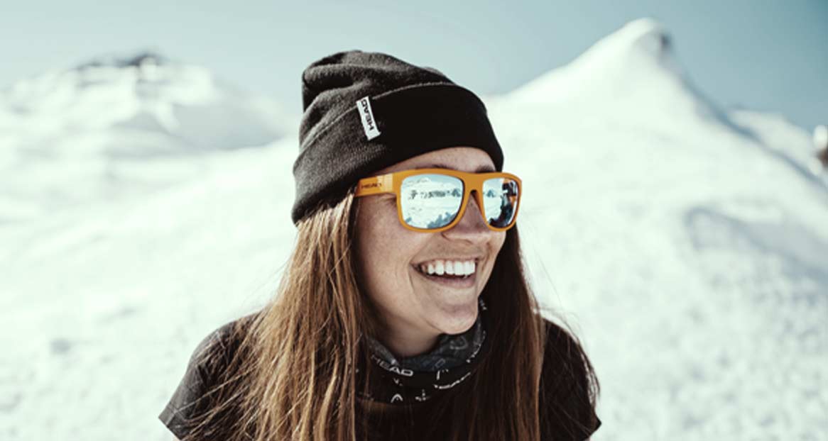 Eine Frau lächelt mit orangefarbener Sonnenbrille vor schneebedeckten Bergen. Sie trägt eine schwarze Mütze.