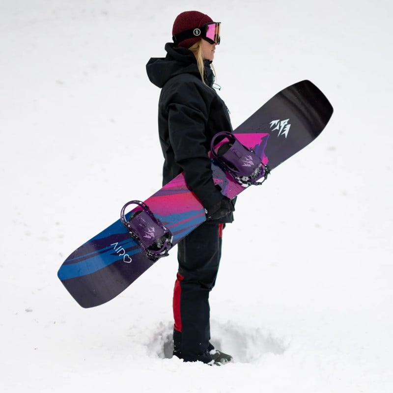 Jones Airheart 2.0 Snowboard Frau hält Jones Airheart 2.0 Snowboard mit farbigem Design im Schnee.