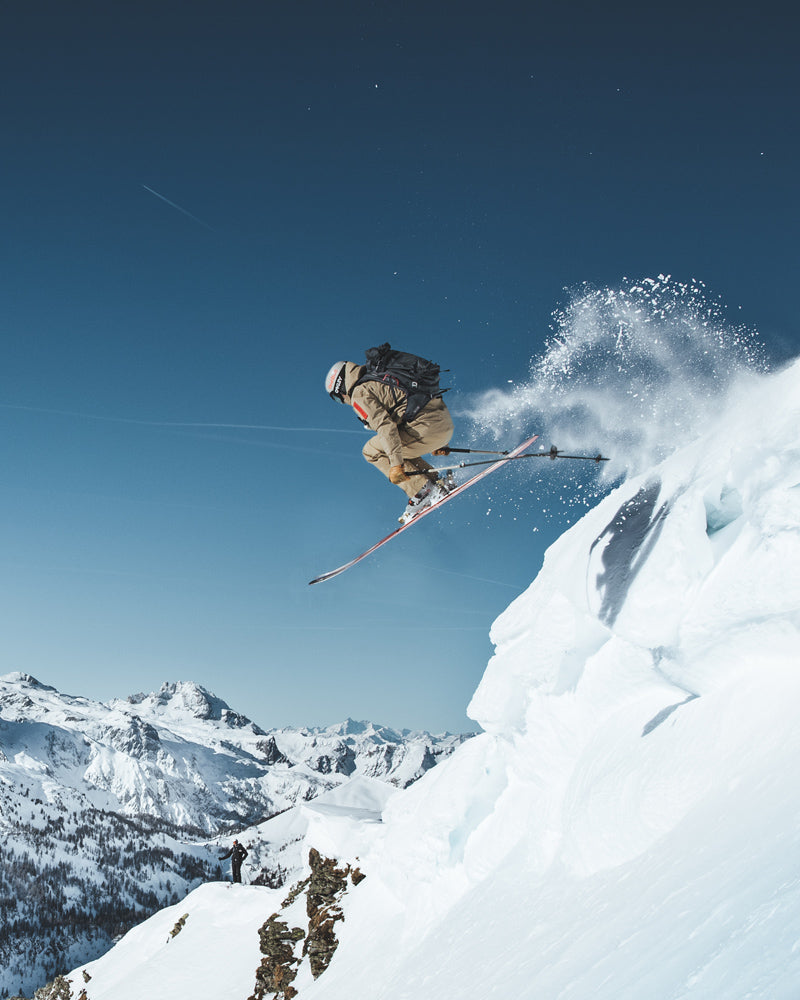 Entdecke die Marke: The Mountain Studio Ein Skifahrer springt von einem schneebedeckten Felsen in den Bergen vor blauem Himmel.