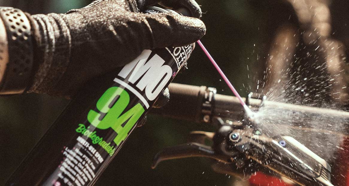 Eine Hand in einem schwarzen Handschuh sprüht mit einer Dose "MO-94" Reinigungsmittel auf ein Fahrrad, wodurch Spritzer entstehen.