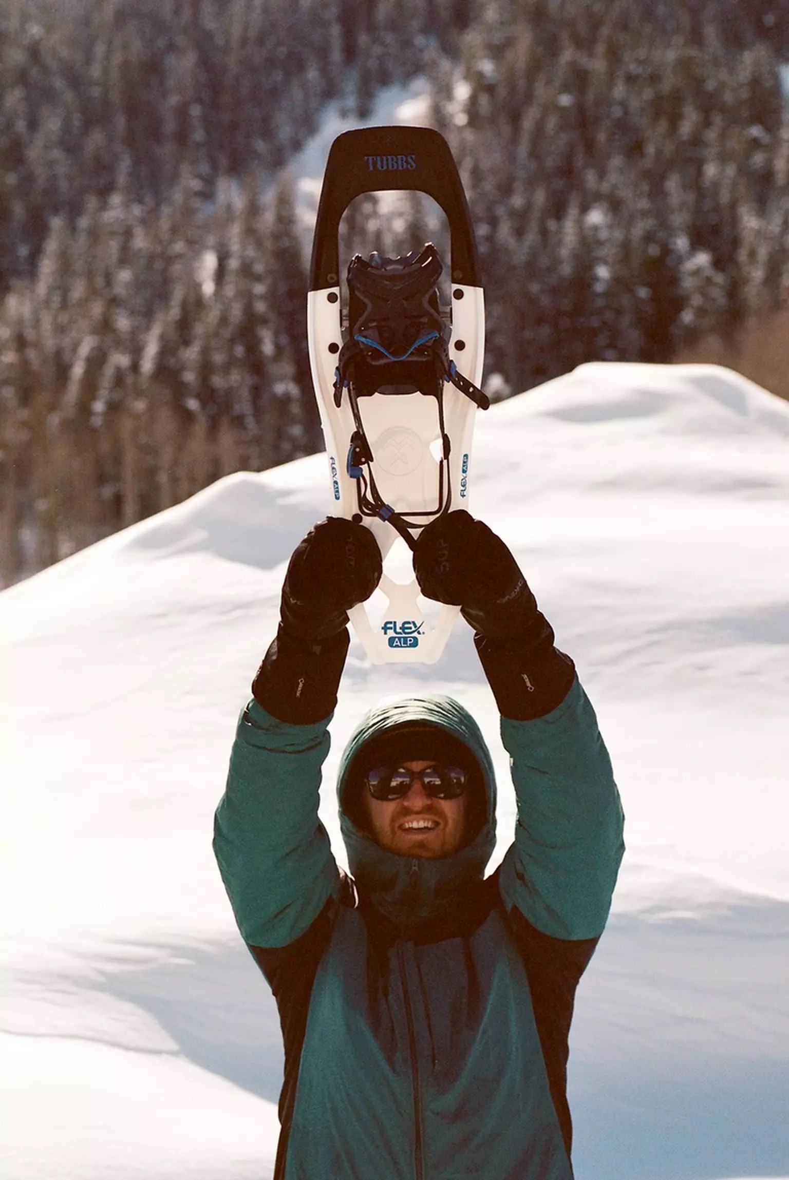 Tubbs Flex Alp 25 Mann hält Tubbs Flex Alp 25 Schneeschuh in Weiß/Blau mit verschneiter Berglandschaft im Hintergrund hoch.