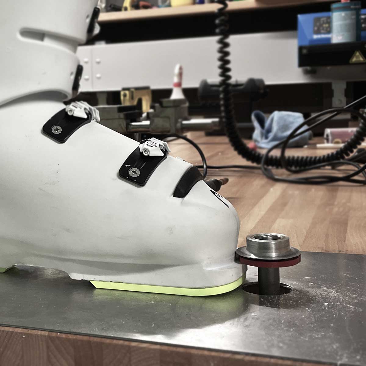 Skischuhe Montage von FIS Lifter Rennplatten Ein weißer Skischuh steht auf einer Metallplatte, mit einer Metallvorrichtung unter dem Schuh. Werkstatthintergrund ist unscharf.