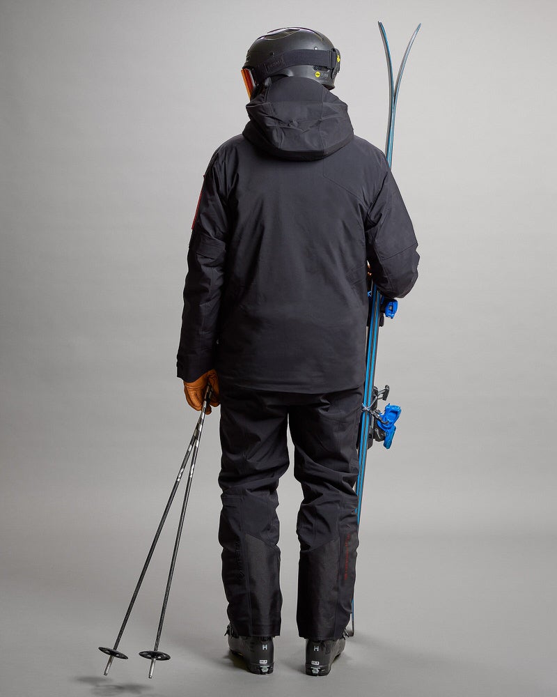 Skifahrer in Gore Tex Person von hinten mit Ski, Stöcken, Helm und schwarzer S-1 Gore Tex 2L Stretch Insulated Jacke und Hose.