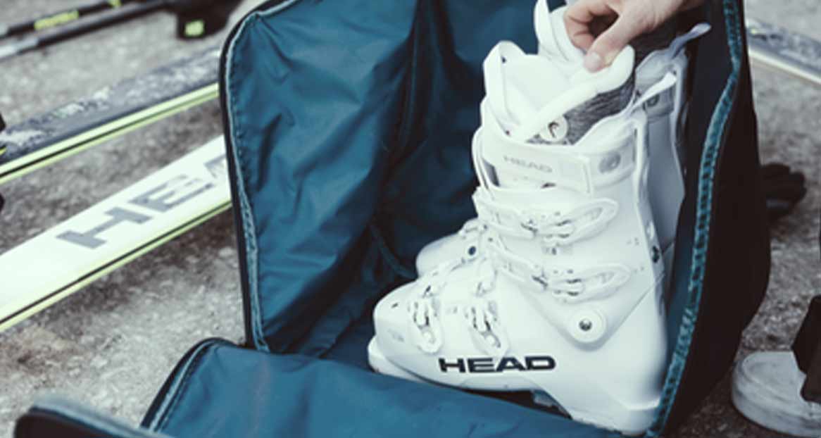 Eine geöffnete Skischuhtasche mit einem weißen Skischuh der Marke HEAD, Skier und Skistöcke liegen daneben.