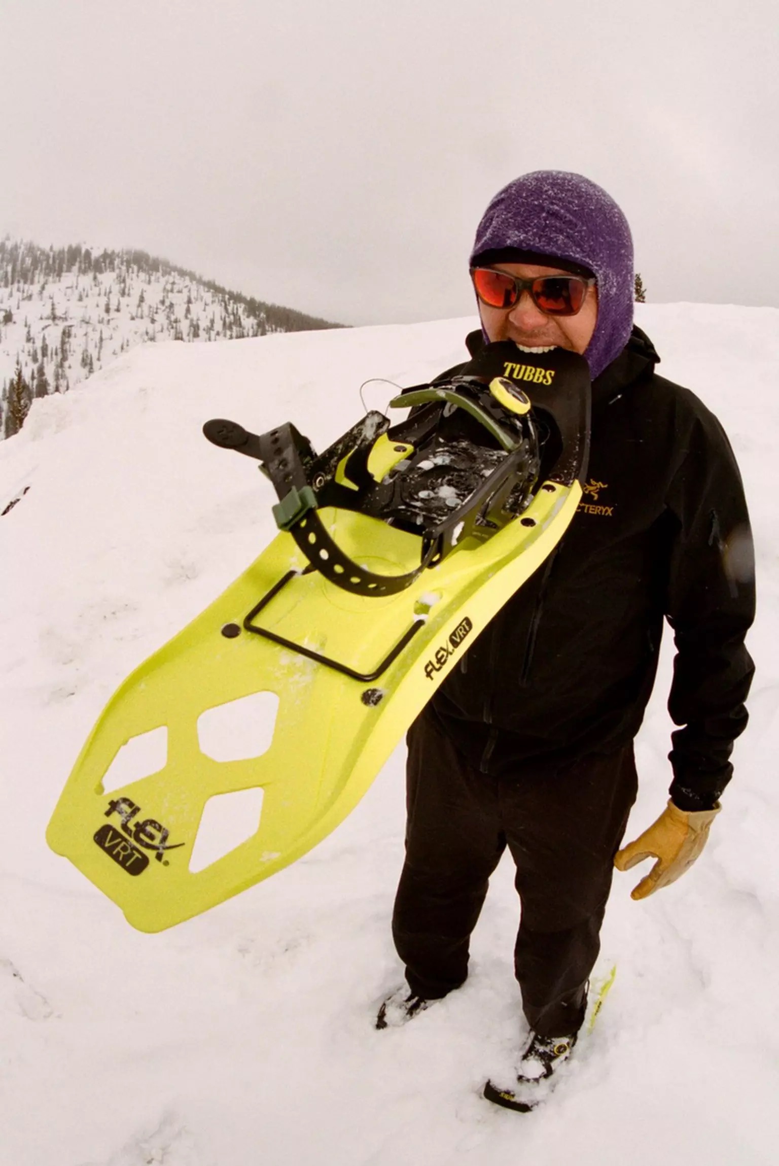Mann mit Schneeschuh Ein Mann hält einen gelben Tubbs Flex VRT Schneeschuh im Mund, steht im Schnee mit Bergen im Hintergrund.