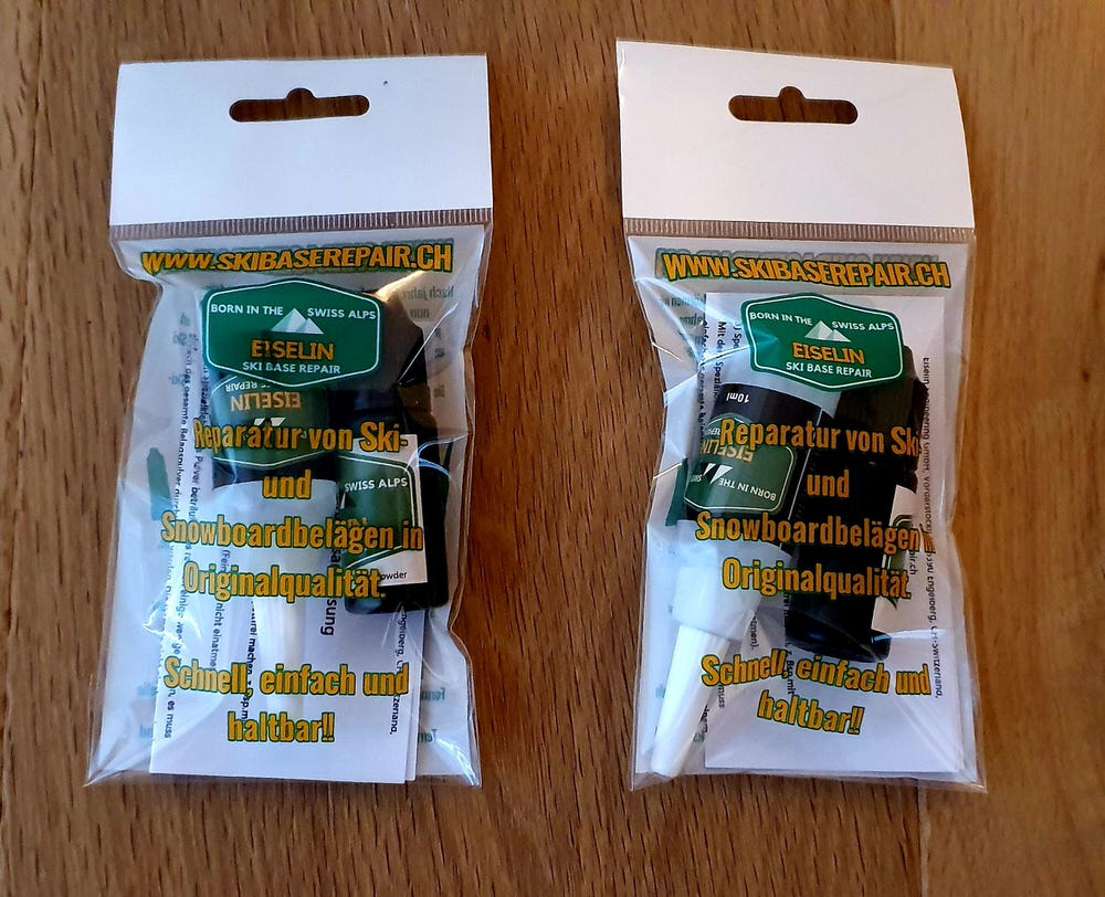 Zwei Ski Base Repair Sets in durchsichtigen Verpackungen auf einer Holzoberfläche. Die Sets enthalten Ski Belagspulver und Kleber zur Reparatur.