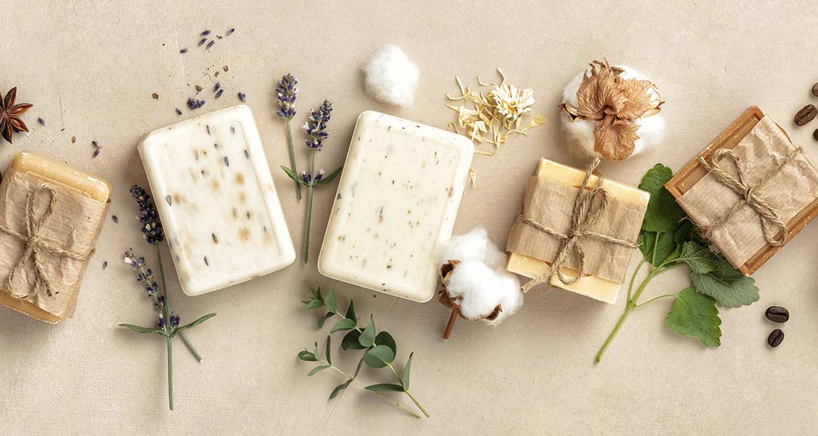 Flach liegende Handseifen, teils verpackt mit Papier und Bindfaden, Lavendelblüten, Baumwolle und Kaffeebohnen auf beigem Grund.
