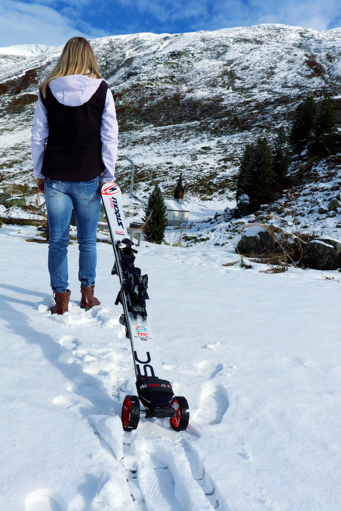 Skifahrerin im Schnee Frau mit Skiern auf Schnee, ausgestattet mit rotem Skimover Traveller Pro mit Rädern. Im Hintergrund ist ein Berg und blauer Himmel sichtbar.