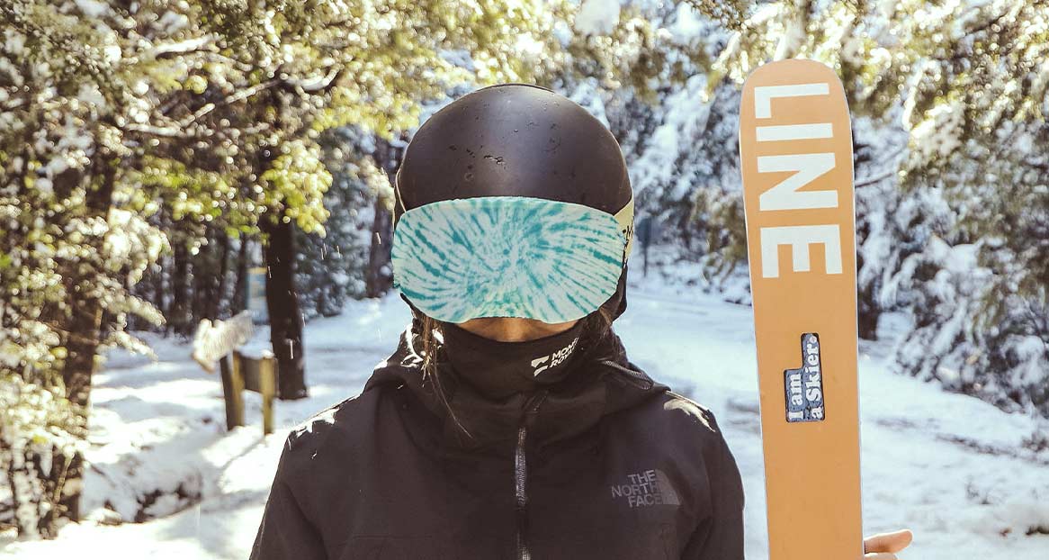 Skifahrer mit Helm und Gogglesoc steht im Schnee. Eine Skispitze mit dem Wort "LINE" ist sichtbar. Bäume mit Schnee im Hintergrund.
