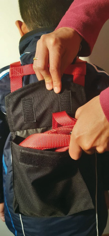 Ski Kindertasche falten Kind mit Skitrainer: Rote Leine wird in eine schwarze Tasche auf dem Rücken verstaut. Elternhände falten die Tasche zu.
