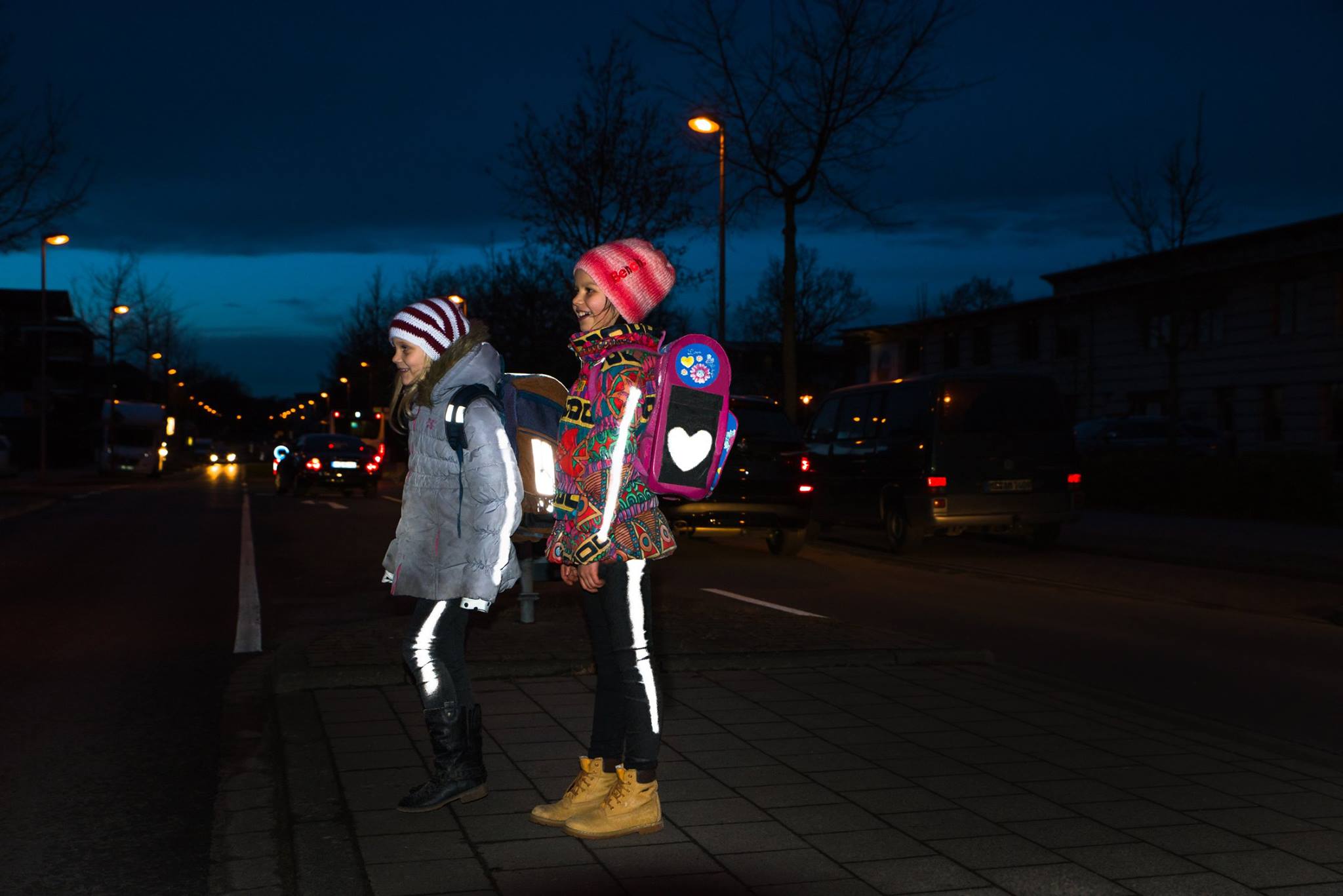 Reflektierender Lightflex-Spray Zwei Mädchen mit reflektierender Kleidung stehen nachts an einer dunklen Straße. Autos sind im Hintergrund zu sehen.