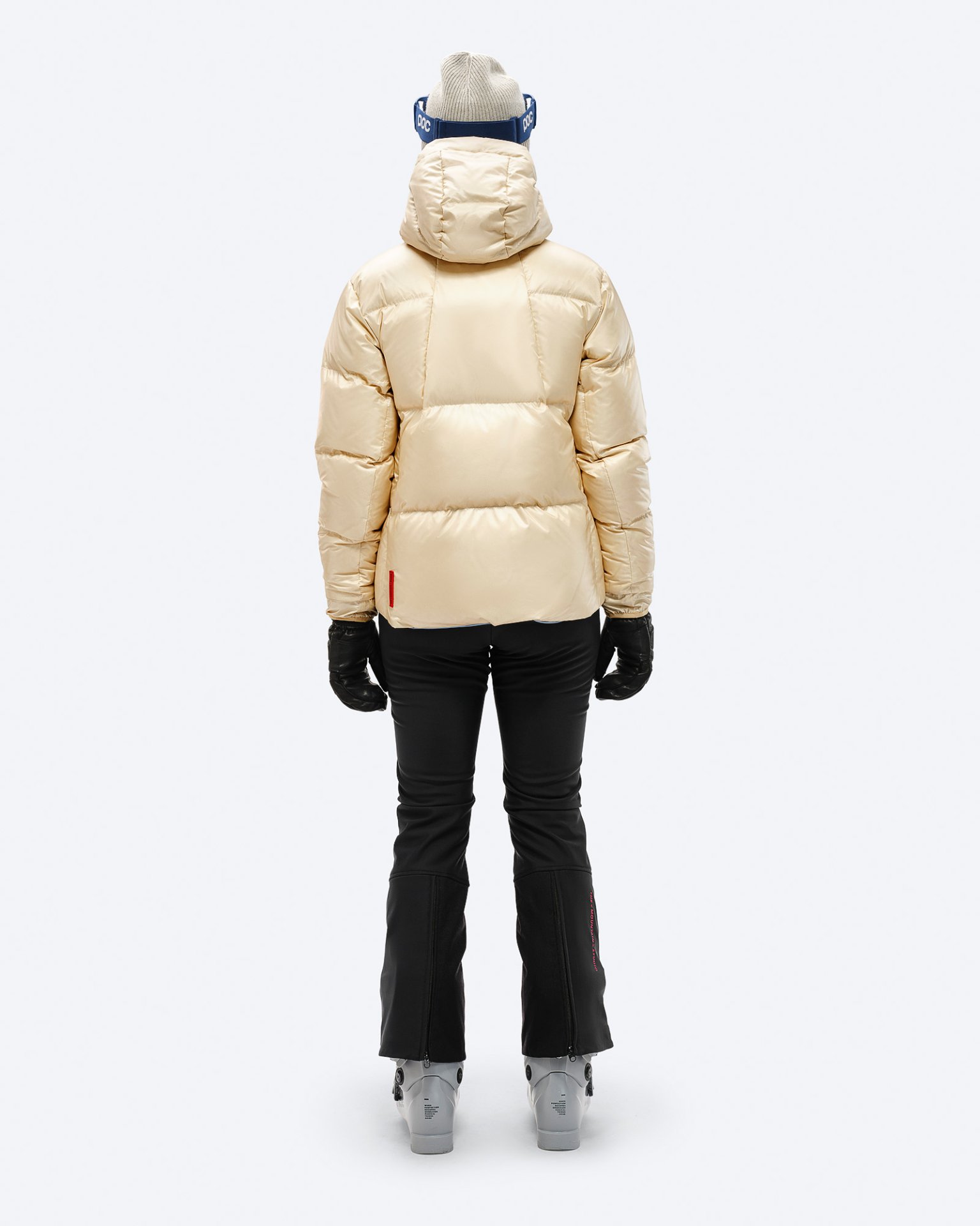 Beige Daunenjacke Rückansicht Rückansicht: Beige Daunenjacke mit Kapuze, schwarze Skihose, Handschuhe und Skischuhe, vor einem weißen Hintergrund.