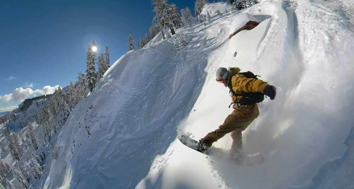 Ein Snowboarder fährt einen steilen, schneebedeckten Hang hinunter, umgeben von Bäumen und unter einem blauen Himmel.