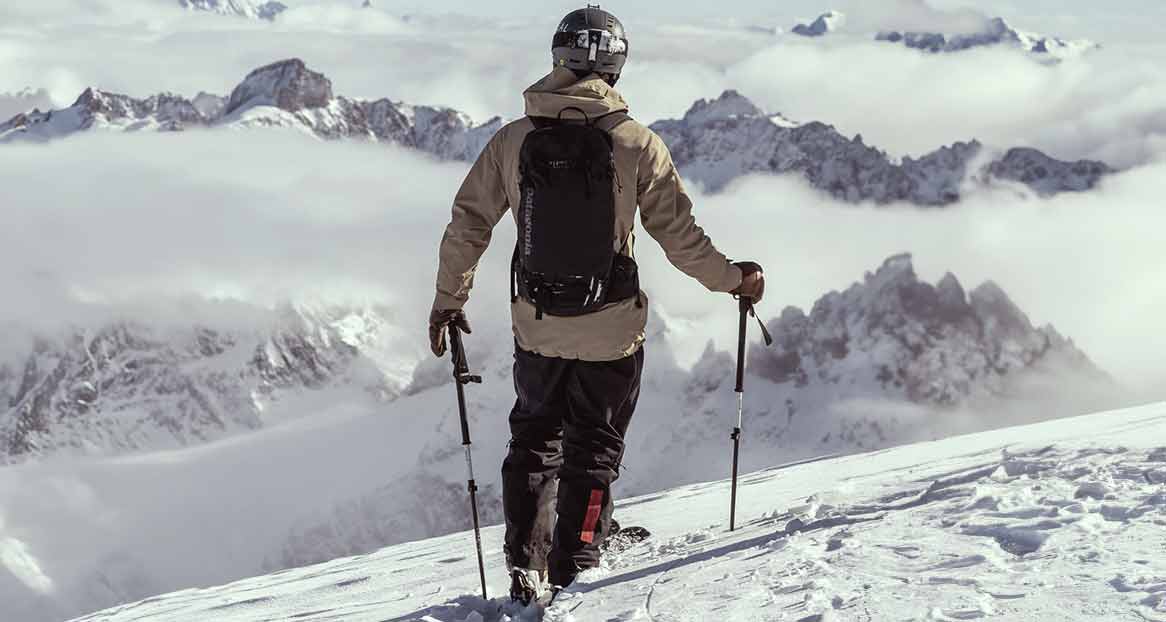 Skifahrer von hinten auf schneebedecktem Hang, mit Stöcken. Im Hintergrund hohe, teils bewölkte Berge.