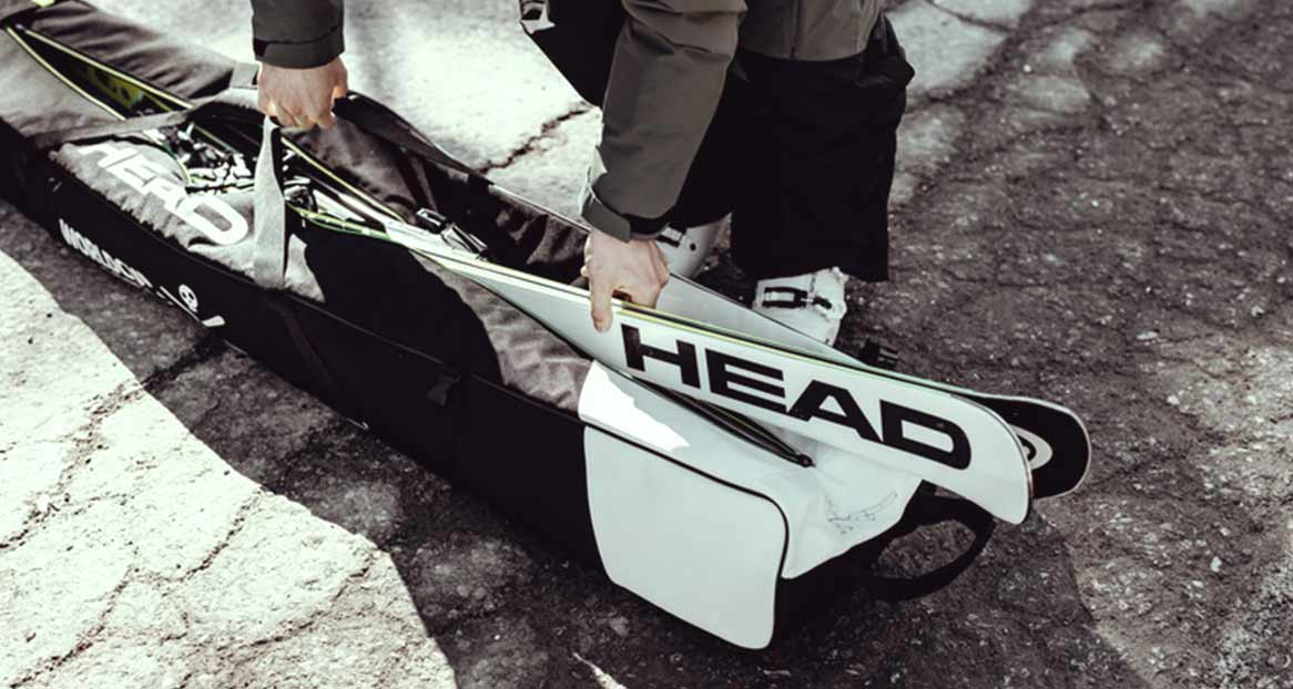 Eine Person packt "Head"-Skier in eine schwarze Skitasche. Die Tasche steht auf rauem Untergrund. Skischuhe sind im Hintergrund sichtbar.
