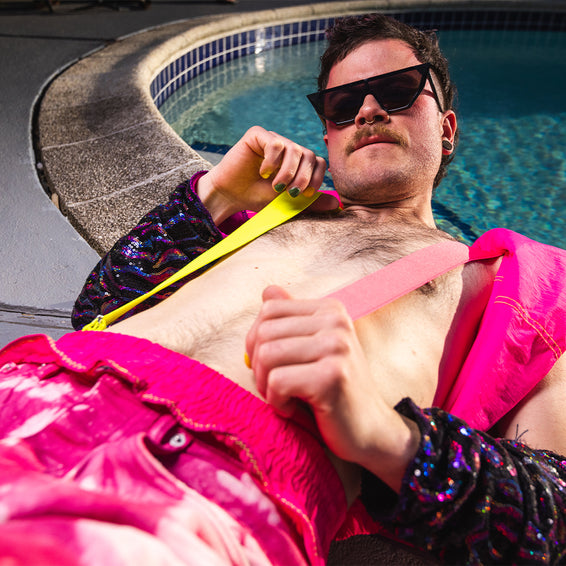 Homme avec lunettes de soleil, torse nu, avec bretelles et pantalon rose, allongé au bord de la piscine.