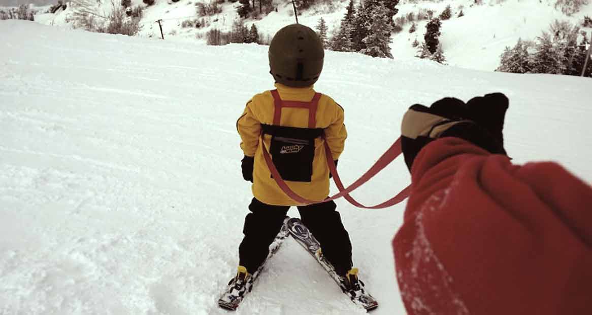 Kind mit Skihelm und gelber Jacke, das von einer Person mit rotem Handschuh an einer roten Leine auf Schnee gezogen wird.
