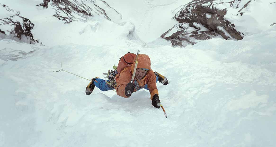 Ein Bergsteiger klettert eine verschneite Bergwand hinauf. Er trägt eine orangefarbene Jacke und blaue Hose und hat Steigeisen an.