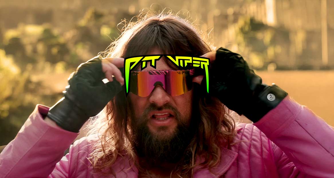 Mann mit Bart setzt Pit Viper Sonnenbrille auf, trägt pinkes Jackett und schwarze Handschuhe vor einem unscharfen Hintergrund.