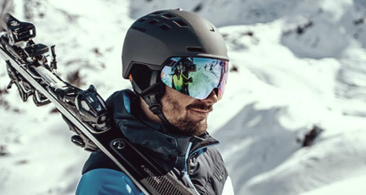 Mann mit Skihelm und reflektierender Brille, Skier über der Schulter, im Hintergrund verschneite Berge.