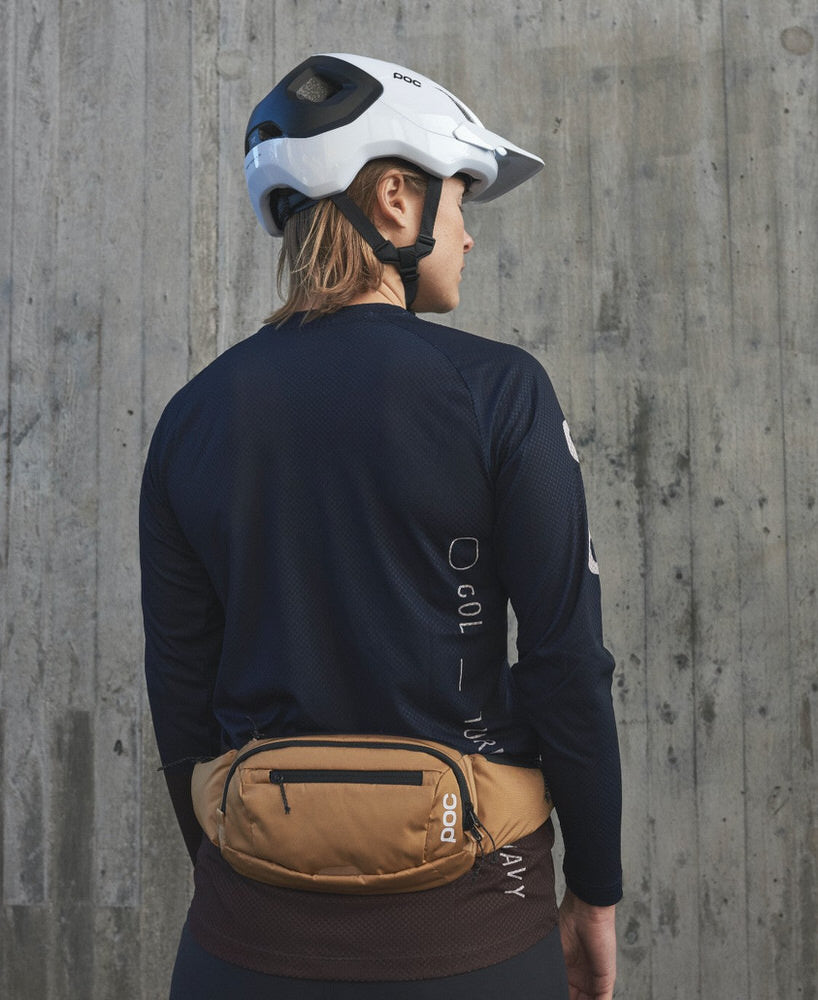 Braune Hüfttasche Tragen Eine Person trägt eine braune POC Lamina Hüfttasche vor einer Betonwand. Sie trägt auch einen Helm und ein langärmeliges Shirt.