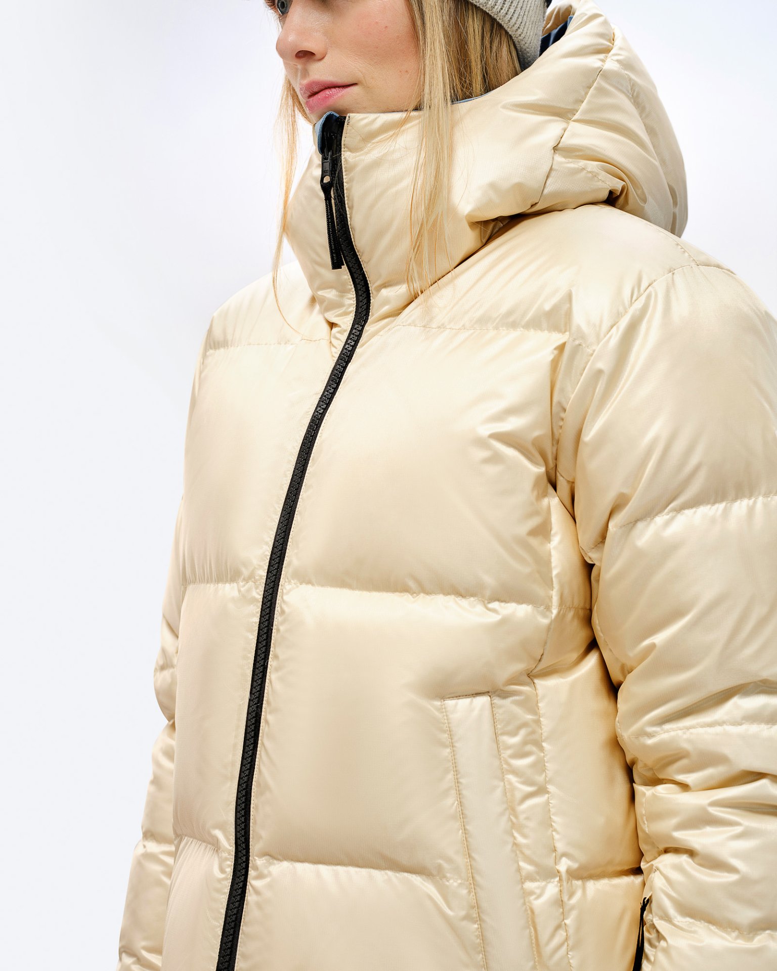 Beige Daunenjacke mit Kapuze Person in glänzender, beiger Daunenjacke mit Kapuze und schwarzem Reißverschluss. Steppjacke, seitliche Ansicht.
