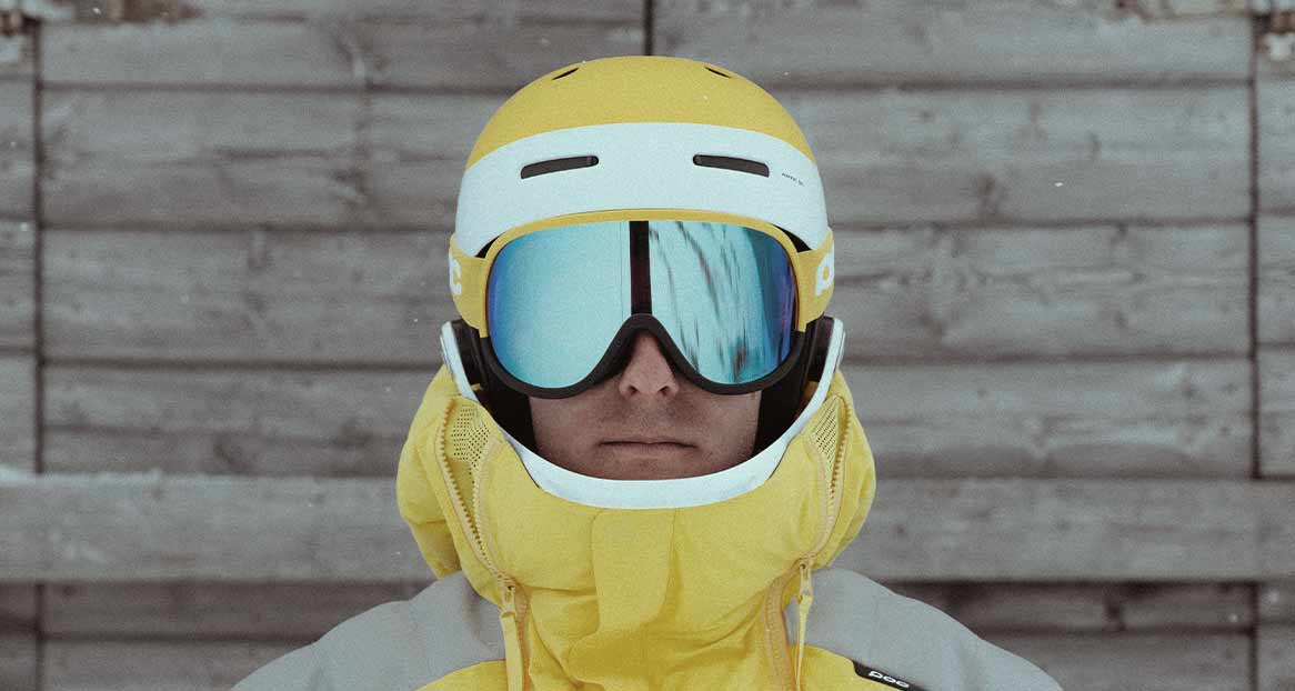 Porträt einer Person mit gelbem Skihelm und blau verspiegelter Skibrille vor einer grauen Holzwand. Gelbe Jacke ist sichtbar.