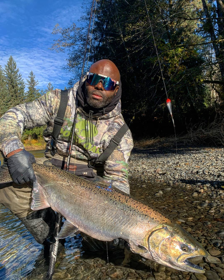Homme portant des lunettes de soleil polarisées tenant un gros poisson dans une rivière, environnement boisé.