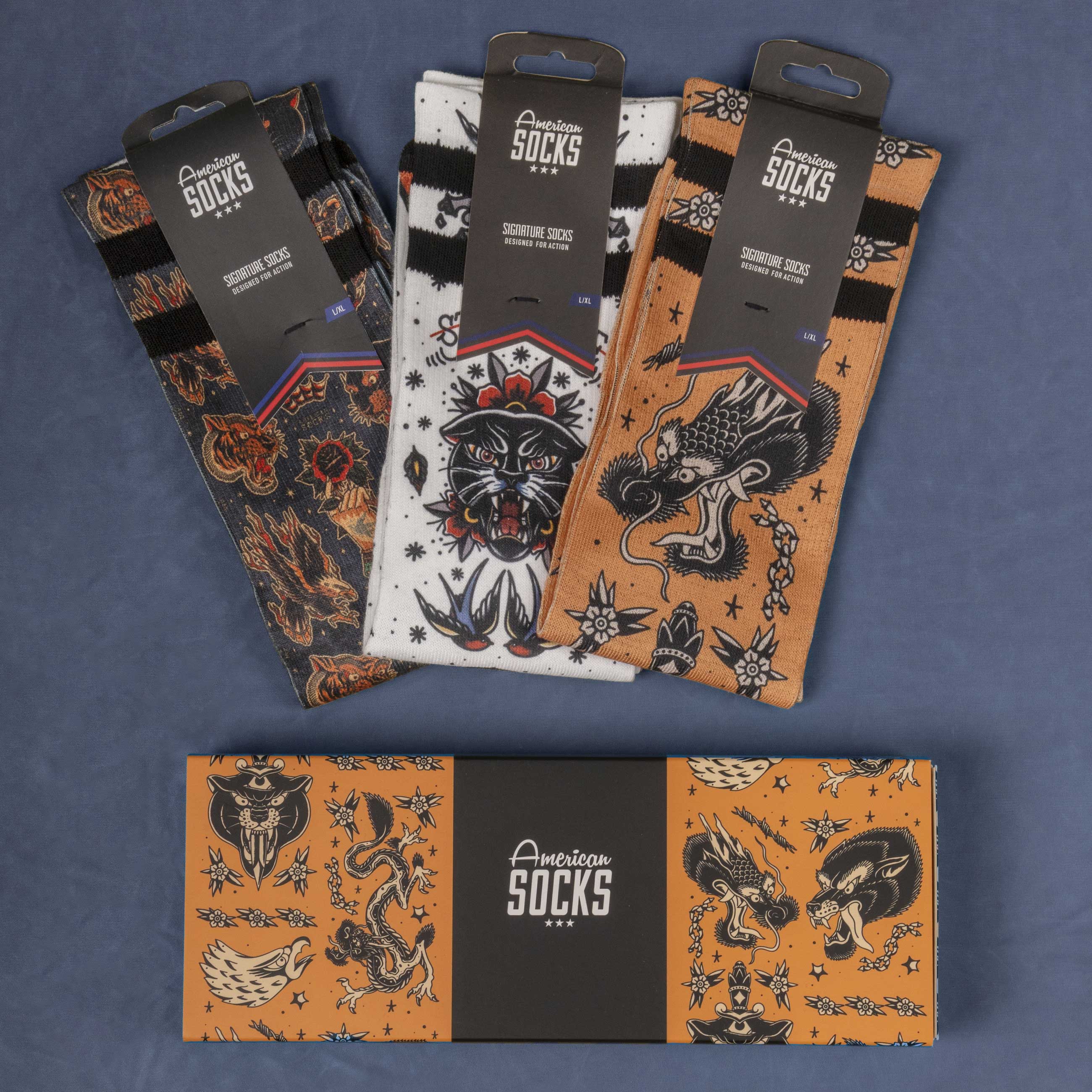 Tattoo-Socken im Geschenkbox Drei verpackte American Socks mit Tattoo-Motiven und eine passende Geschenkbox liegen auf einem blauen Untergrund.