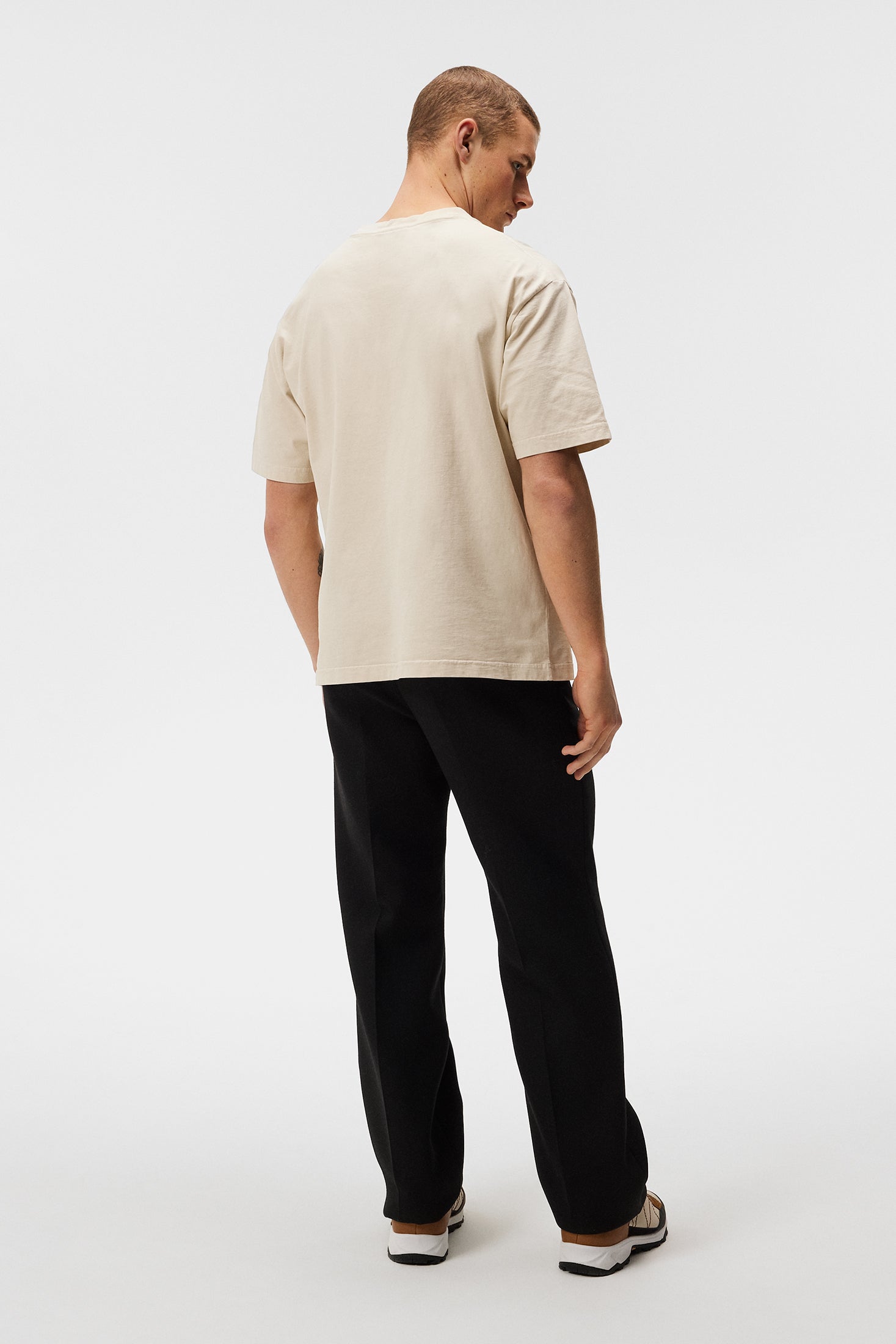 Ein Mann trägt ein Oyster Gray Walsh Boxy Print Tee, kombiniert mit schwarzen Hosen, von hinten auf einem weißen Hintergrund.