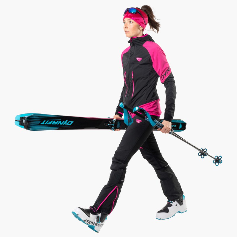 Skifahrerin mit Ausrüstung Frau in Skikleidung mit Blacklight 88 Ski unter dem Arm und Skistöcken in der Hand, geht vorwärts.