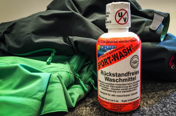 Sport-Wash - Waschmittel | ski-shop.ch Grüne und graue Jacke, neben einer Flasche Sportwaschmittel auf grauem Untergrund.