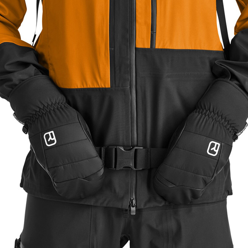 Detailaufnahme: Schwarze Ortovox Freeride Cozy Fäustlinge vor einer Winterjacke mit Gürtel.