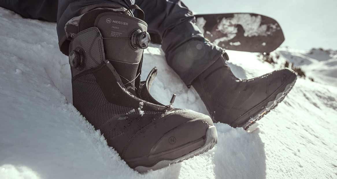 Schwarze Snowboardschuhe der Marke Nidecker im Schnee. Im Hintergrund ist ein Snowboard und eine schneebedeckte Berglandschaft zu sehen.