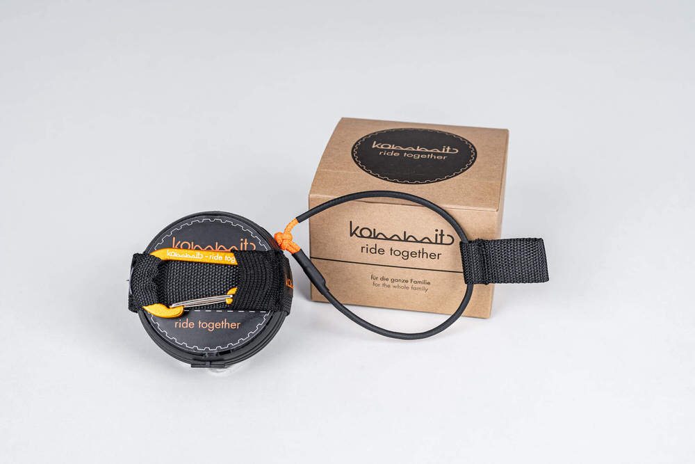Zugsystem mit Karabiner Das kommit Zugsystem: Eine schwarze Spule mit Gurtband und Karabiner liegt neben einer braunen Schachtel. Schwarzes Seil verbunden.