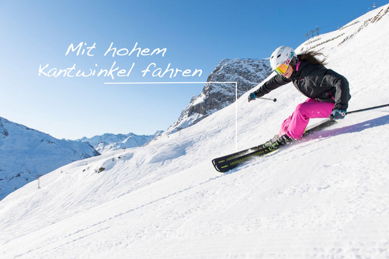 Tipps, wie sie als guter Skifahrer noch besser werden. Skifahrerin mit pinker Hose auf schneebedeckter Piste vor Bergkulisse unter blauem Himmel.