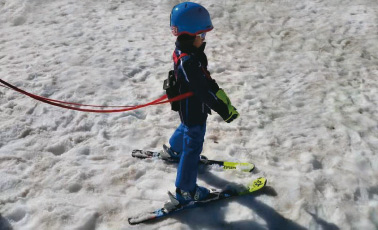 Ski Trainer Kind Kind mit Ski Trainer auf Schnee. Das Kind trägt einen blauen Helm und wird mit Leinen geführt.