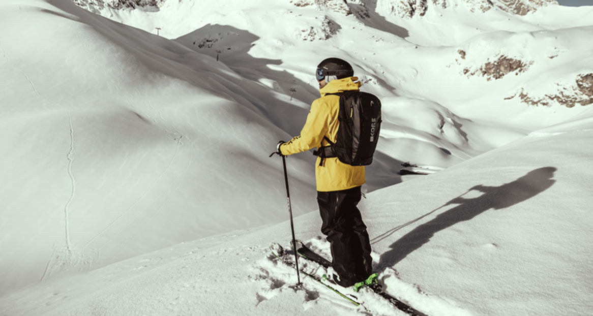 Ein Skifahrer mit gelber Jacke und Rucksack steht im Schnee, mit Blick auf schneebedeckte Berge.