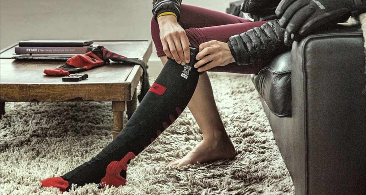 Person sitzt auf Couch und reguliert eine beheizbare, schwarze Socke mit roter Spitze. Auf dem Tisch liegen Bücher und eine zweite Socke.
